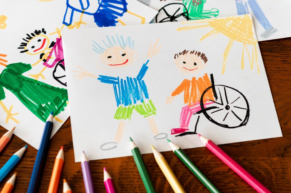 Kinderzeichnung von einem lachenden Kind und einem lachenden Kind im Rollstuhl mit Buntstiften herumliegend
