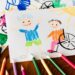 Kinderzeichnung von einem lachenden Kind und einem lachenden Kind im Rollstuhl mit Buntstiften herumliegend